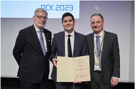 Drei Männer in Anzügen, einer hält eine Urkunde. Im Hintergrund das Logo "DCK 2023".