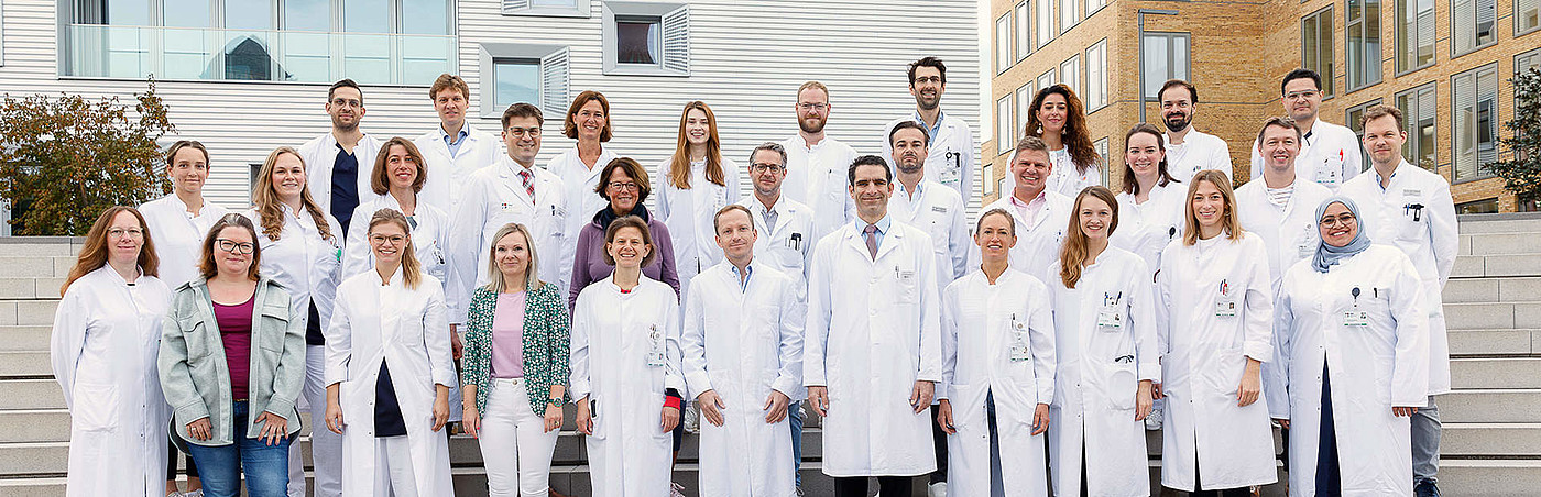 Chirurgie Gruppenfoto von 27 Ärzten und Ärztinnen in weißen Kitteln, die auf einer Treppe vor einem modernen Gebäude stehen.