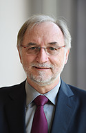 Ein älterer Mann mit Brille, Anzug und roter Krawatte lächelt vor einem verschwommenen Hintergrund.