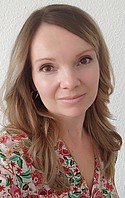 Eine Frau mit schulterlangem Haar lächelt in die Kamera. Sie trägt eine geblümte Bluse und steht vor einer weißen Wand.