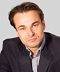 Mann in dunklem Anzug und gestreiftem Hemd schaut in die Kamera vor grauem Hintergrund.