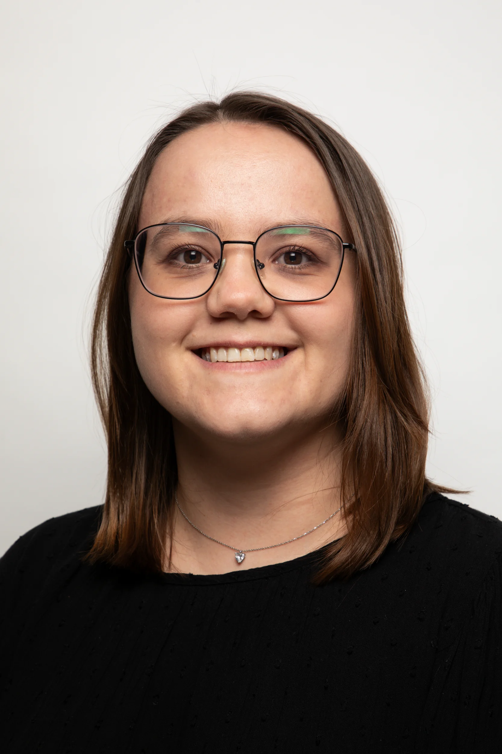 Porträt einer lächelnden Person mit Brille und braunen Haaren, die ein schwarzes Oberteil und eine Kette trägt.