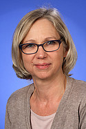 Eine Frau mit blonden Haaren und Brille vor blauem Hintergrund. Sie trägt einen beigen Pullover und schaut freundlich.