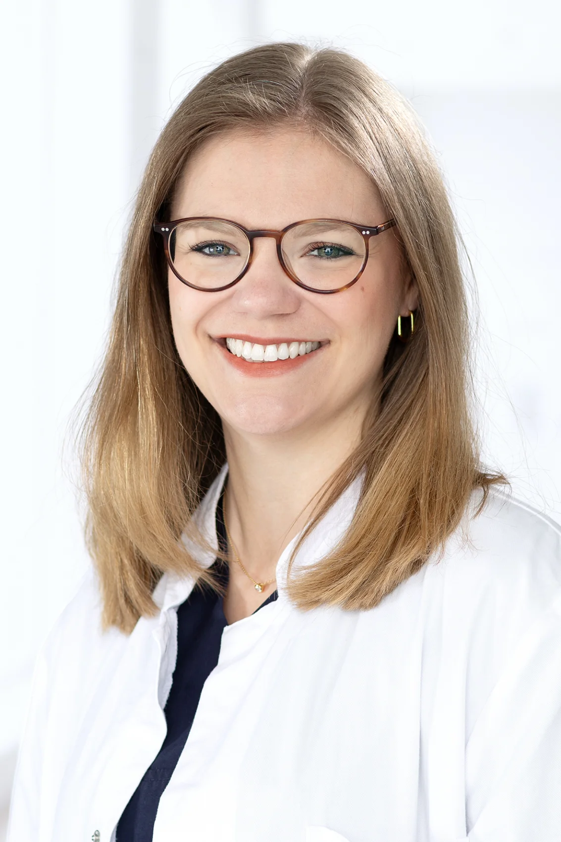 Frau mit Brille und weißen Kittel lächelt in die Kamera. Hintergrund unscharf.