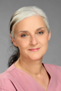 Porträt einer Frau mit grauen Haaren, die ein rosafarbenes Hemd trägt. Sie lächelt leicht vor einem grauen Hintergrund.
