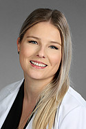Frau mit langen, blonden Haaren und weißem Kittel lächelt vor neutralem grauen Hintergrund.