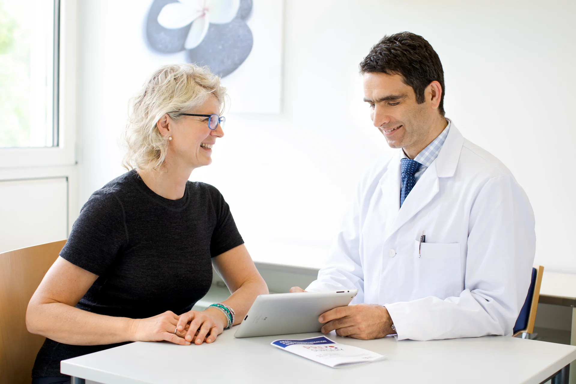 Ärztin und Patientin sitzen an einem Tisch, lächeln und betrachten gemeinsam ein Tablet.