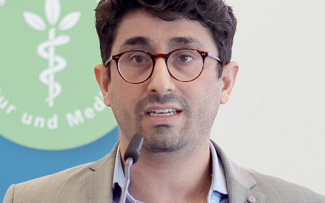 Mann mit Brille spricht in Mikrofon, Hintergrund zeigt ein Logo mit grünem Kreis und Pflanzensymbol.