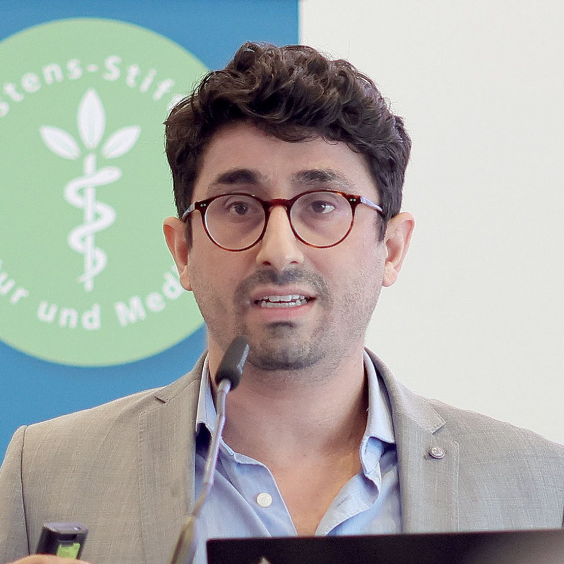 Mann mit Brille spricht in Mikrofon, Hintergrund zeigt ein Logo mit grünem Kreis und Pflanzensymbol.