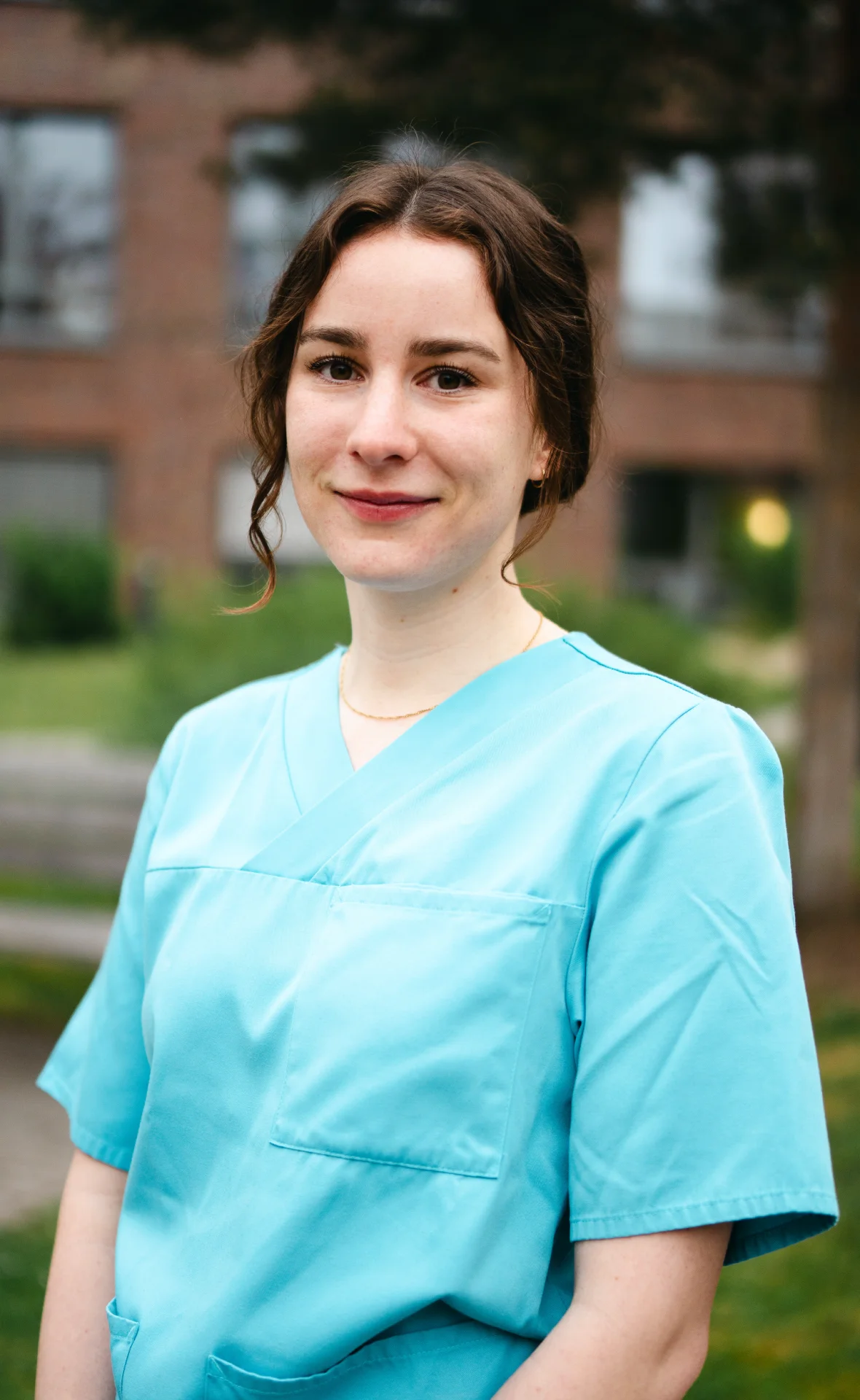 Frau in blauer Pflegeuniform lächelt im Freien vor einem unscharfen Hintergrund.