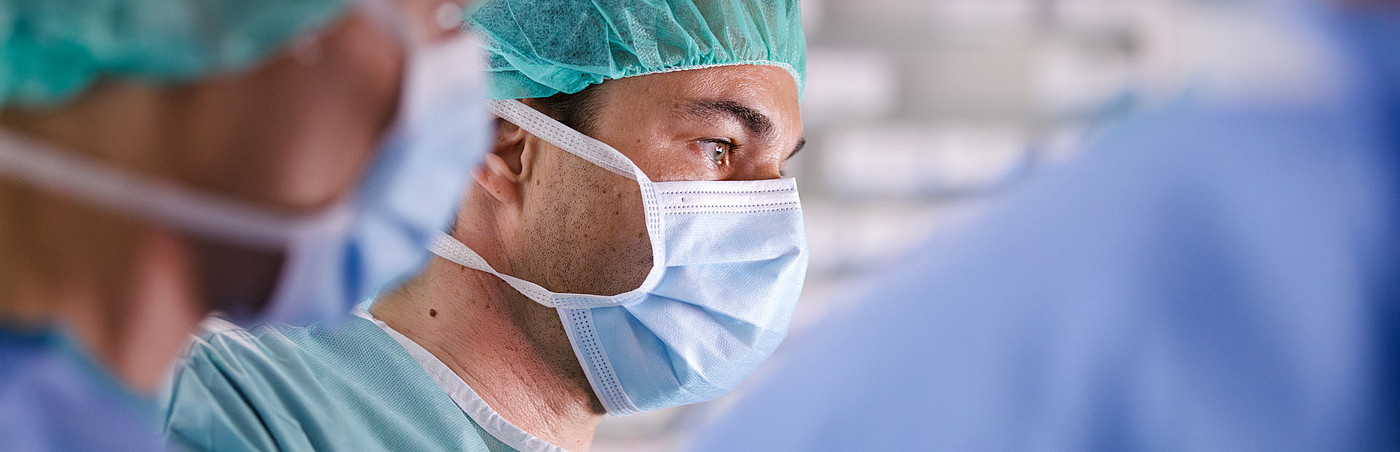 Chirurg mit Mundschutz und Haube konzentriert bei einer Operation. Umgeben von anderen in OP-Kleidung.
