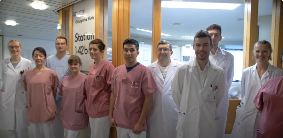 Medizinisches Team in weißer und rosa Arbeitskleidung steht vor einer Stationstür in einer chirurgischen Klinik.