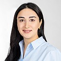 Portrait einer lächelnden Frau mit dunklem Haar in blauem Hemd vor hellem Hintergrund.