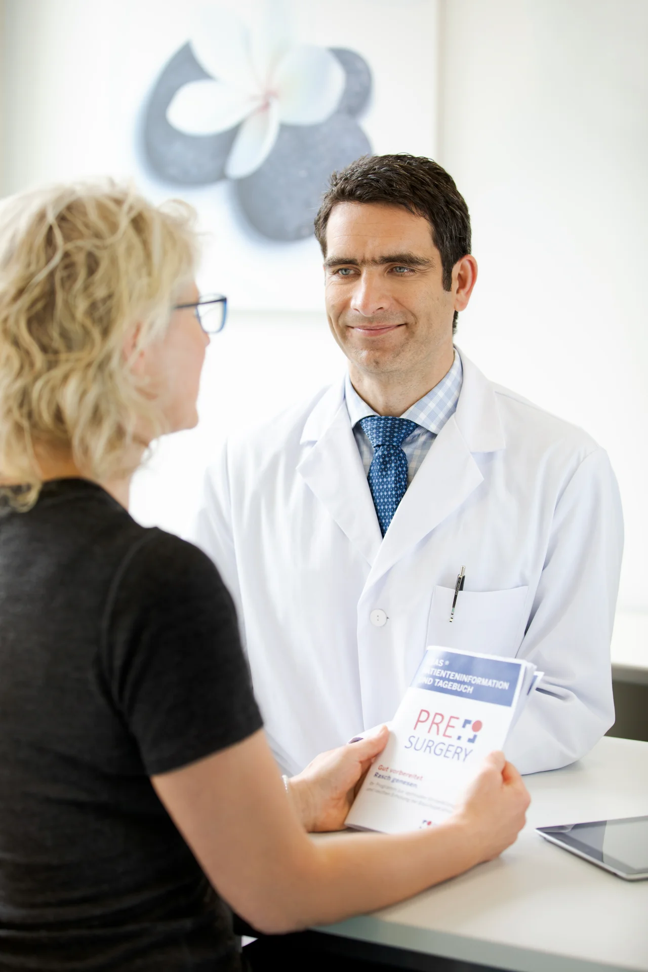 Arzt im Gespräch mit Patientin, hält Broschüre mit der Aufschrift "Pre-Surgery", beide lächeln.