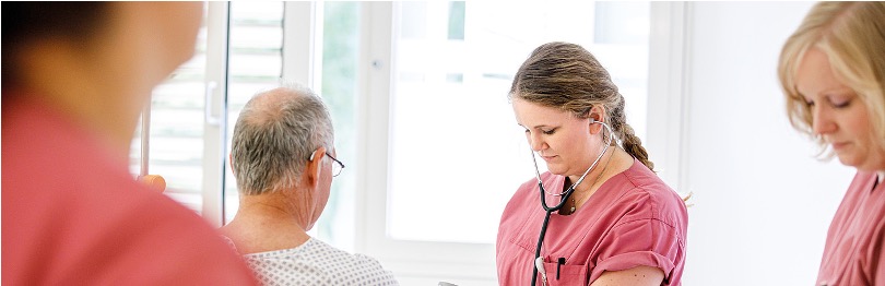 Krankenpflegerin in rosa Kittel untersucht Patienten mit Stethoskop, während zwei weitere Pflegekräfte anwesend sind.